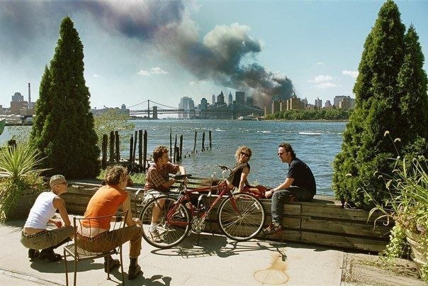 США. Бруклин, Нью-Йорк. 11 сентября 2001. Молодые люди отдыхают во время перерыва на обед, в то время как огромный столб дыма поднимается от нижнего Манхэттена после нападения на Всемирный торговый центр.